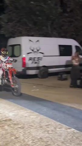 The video begins with a blurred shot of a person on a dirt bike at night, wearing a red and white racing suit and helmet. The camera then pans up to show the person and the dirt bike in mid-air, performing a jump against a dark night sky with some lights visible in the distance. The bike is red and white. The person appears to be in a white suit with red accents. The jump takes place over what appears to be an inflatable landing ramp, which is visible in the lower portion of the frame. The video then shows the ramp from a different angle, revealing a large black inflatable structure and sound equipment. The ground appears to be dirt or sand. The overall scene depicts a nighttime motocross stunt performance.