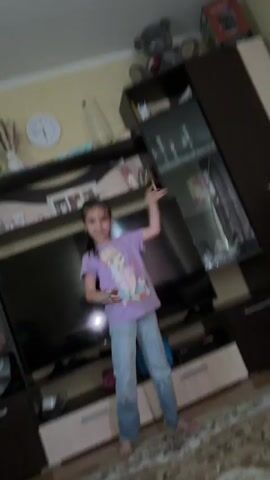 In this media file, a young girl is filmed in a living room setting. She is the primary subject, standing barefoot on a patterned rug in front of a large television. She is wearing a light purple t-shirt with a graphic print and light blue jeans. The girl is actively dancing and interacting with the camera, with her right arm raised and her hand in a gesture towards the viewer. Her facial expression suggests a playful and engaged demeanor.

The background reveals a typical home environment. A large television is mounted on a dark wood entertainment unit. Above the television, shelves are visible, displaying various decorative items, including a vase with dried flowers, a framed photograph, and a wall clock with Roman numerals. To the right of the television, a tall, dark wood cabinet with glass doors showcases more items. A teddy bear is perched on the top shelf of this cabinet. The overall lighting suggests an indoor setting, possibly during the day, as there are no strong indications of artificial lighting being the sole source.

The activity captured is a spontaneous dance or performance by the child. Her movements are energetic and her gestures are directed at the camera, implying she is aware of being filmed and is participating in the recording. The setting appears to be a comfortable, lived-in home space, likely in Tambov, Russia, given the provided location context. There are no discernible external weather conditions to comment on. The emotions conveyed are primarily positive, with the child appearing happy and animated. No visible text is present within the frame.
