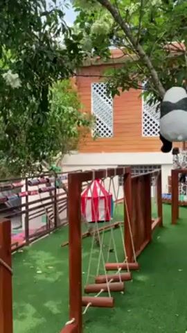 Here's a concise description of the video:

The video shows an outdoor play area, likely in a daycare or preschool setting. The area is covered in artificial grass and includes wooden structures with rope ladders and climbing walls.  A small red and white striped tent is also visible. A stuffed panda toy hangs from a tree branch.  The background includes a light brown building with white-lattice windows. The sky is partially visible with some clouds. A child is briefly seen running in the background.