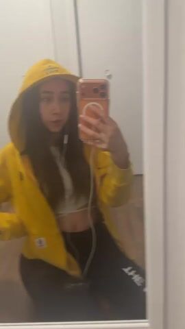 A young woman with long brown hair is shown in a selfie in front of a mirror. She is wearing a yellow hooded sweatshirt with a white cropped top underneath, and black leggings with white text down the side. Her phone has an orange-colored case. She has earphones in. The background shows a white door and what appears to be a wooden floor. At first she looks serious, but then she smiles.