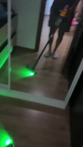 A person is in a room, holding a cordless vacuum cleaner that has a green light on the end of the cleaning head. The person is using the vacuum cleaner to clean the floor, and the green light illuminates the floor as they vacuum. The person is wearing shorts and a t-shirt, and is standing in front of a mirror, so their reflection is visible. The room has wooden floors and appears to be a bedroom, with a dresser and a door visible in the background.