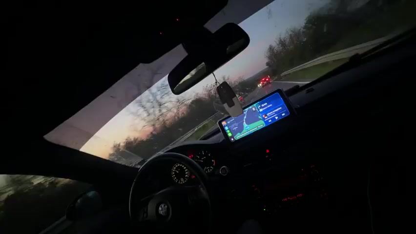 The view from inside a car at dusk shows a road ahead with another car in front. The interior of the car is dark, but the dashboard, steering wheel, and a navigation screen are visible.  The sky is visible through the windshield, showing a faint glow of sunset.