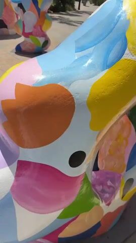 The video shows a close-up, then a wider view, of colorful, abstract sculptures situated on a paved walkway in an outdoor area. The sculptures are painted with a variety of bright, organic shapes in hues of pink, blue, yellow, orange, purple, and green. In the background, there are palm trees, buildings with balconies and awnings, and a clear blue sky. The paved walkway is made of interlocking stones. One sculpture resembles an archway with pointed elements at the top, while another is a smaller, amorphous shape. The camera pans slowly across the scene, capturing the details of the artwork and its surroundings. No people are clearly visible interacting with the sculptures. The overall impression is one of modern outdoor art in a sunny, possibly Mediterranean, environment.