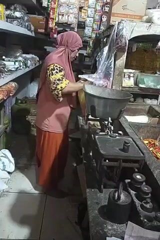 A woman wearing a pink hijab and a pink shirt with a leopard print sleeve is standing in what appears to be a store. She is holding a yellow plastic bag and placing it onto a large metal scale. In the background, there are shelves filled with various products and a green gas tank. The scale has weights on it, and there is a large metal bowl on top of the scale. The woman is looking down at what she is doing.