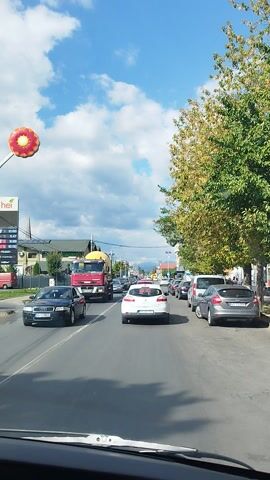 This media file captures a view from inside a vehicle driving on a road in Curtea de Argeș, Romania. The scene depicts a traffic jam on a sunny day with scattered clouds. The road is lined with buildings, businesses, and trees with some autumn foliage.

In the foreground, the dashboard of the vehicle is visible, suggesting the perspective of a driver or passenger. Several cars are on the road, including a red cement truck on the left, a white hatchback in the center, and various other vehicles of different colors and types. A silver sedan is parked on the right side of the road, and further down the street, more cars are visible.

A large, red and yellow inflatable flower hangs in the sky on the left side of the frame. On a building to the left, there's a sign with text that appears to be related to fuel prices, showing numbers like "0.58", "0.47", and "0.48". Another sign on the same building reads "Cazare", which means "Accommodation" in Romanian.

The overall atmosphere is one of a typical urban commute, with the bustle of traffic under a bright, partly cloudy sky. The presence of autumn colors in the trees hints at the season.