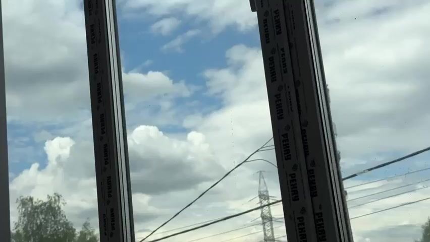 The video shows a view through a window frame. The frame is white and has text printed along the edges. The text appears to be repeating the word "PENRY" multiple times. Outside the window, there is a blue sky with fluffy white clouds. In the distance, there are some trees and a tall transmission tower with wires. The lighting suggests it is daytime, likely overcast due to the clouds.