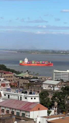 A large orange cargo ship is sailing on a calm body of water. The ship is filled with colorful shipping containers. In the foreground, there is a city with several buildings. The buildings are a mix of architectural styles and are close together. The sky is blue with some white clouds. The overall scene appears to be a harbor or a coastal city.