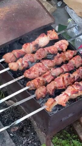 This is a close-up, high-angle shot of skewers of meat cooking over charcoal in a portable grill. The meat is a light pinkish-red color and appears to be well-marbled. The skewers are arranged in rows on the grill, and the meat is visibly sizzling and browning. Smoke is rising from the grill, and the coals below are glowing red. In the background, there is a crumpled brown paper bag, a clear plastic bottle, and a red and white object that may be a can or bottle label. The grill itself is made of dark metal and appears to be old and somewhat rusted. There are also some green plants and a brick visible around the grill, suggesting an outdoor setting. The overall impression is of a casual outdoor cooking scene.