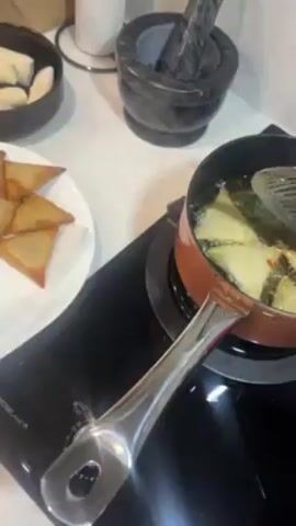 The video shows samosas being deep fried in a small orange pot filled with oil. A metal slotted spoon is used to flip and stir the samosas. A plate with several golden-brown fried samosas on a paper towel is beside the pot. To the upper left of the plate is a small black bowl containing uncooked samosas. Behind the pot is a black mortar and pestle, and a white electric kettle. The cooking surface is a black, glass-top stove.