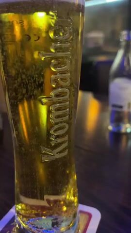A tall glass of Krombacher beer sits on a coaster. The beer is a golden color with many small bubbles rising to the top, where a thin layer of foam has formed. The glass has the Krombacher logo embossed on it. The background is blurred, but appears to be a dimly lit bar or restaurant with other bottles and glasses visible. The lighting is warm and yellow, reflecting off the beer.