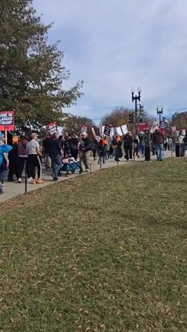 A crowd of people is walking along a paved path in front of a grassy area, carrying signs. Many of the signs appear to be anti-Trump messages, with one specifically stating "Trump Must Go Now!". There is also a large inflatable shark character in the crowd. In the background, the Washington Monument is visible. The weather appears to be partly cloudy. The crowd has a diverse composition, with people of different ages and ethnicities. One person is dressed as a clown. The overall setting suggests a political protest or demonstration.