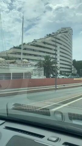 The video shows a view from a vehicle traveling on a road. On the left is a large, curved white building with many balconies. A palm tree is in front of the building. A white truck is driving on the road. In the distance, there are more buildings and trees. The sky is cloudy.
