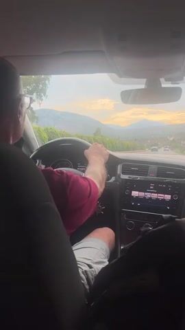 A person wearing glasses and a maroon t-shirt is driving a car. The driver's hands are on the steering wheel. The car's dashboard is visible, with a screen displaying music information. Outside the car, a scenic landscape of hills and mountains can be seen under a cloudy sky with hints of sunset colors. The image appears to be taken from the back seat of the car, looking forward.
