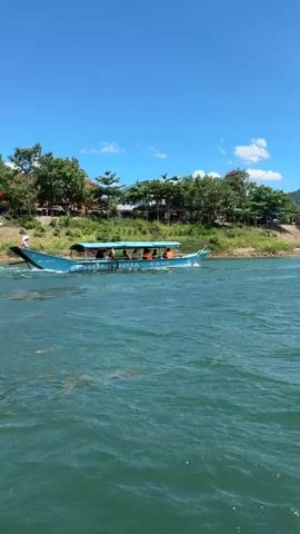 The video shows a boat traveling on a body of water. The boat is blue and has several people on it. The water is a greenish-blue color and appears to be moving. There is a shoreline in the background with trees and some buildings. The sky is blue with some clouds. The camera appears to be on another boat, as indicated by the edge of the boat in the lower right corner of the frame. The video is shaky, suggesting it was filmed while moving.