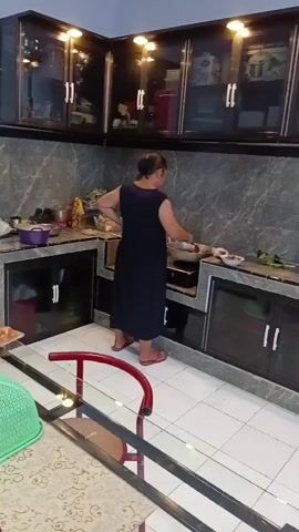 A woman in a dark blue dress and red sandals stands in a kitchen and stirs food in a pan on the stove. She is facing away from the camera. The kitchen has dark cabinets with glass doors and a gray marbled countertop. A white tiled floor is visible. In the foreground, a red chair and a green basket are partially visible.