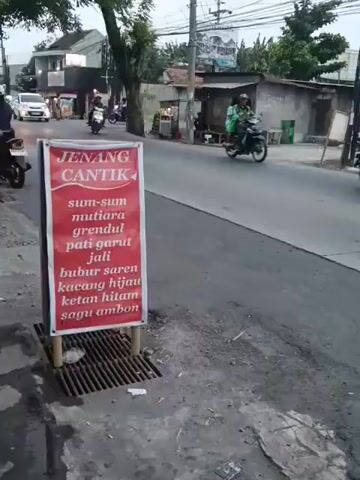 A red sign with white text is placed on the side of a road. The sign reads "JENANG CANTIK" at the top in large white letters, followed by a list of food items in smaller white text. The food items include "sum-sum mutiara", "grendul pati garut", "jali", "bubur saren", "kacang hijau", "ketan hitam", and "sagu ambon". The sign is supported by two bamboo poles. In the background, traffic moves along the road. A white car is seen driving away from the camera on the left side of the road. A motorcycle with two people on it is also seen riding on the road. A black truck is driving past the sign. The environment appears to be an urban or suburban street with buildings and trees visible. The ground is asphalt and dirt, with a grate on the side of the road.