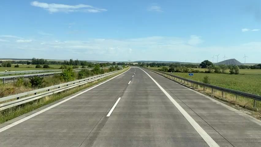 The video shows a forward-facing view from a vehicle traveling on a highway. The highway is paved with concrete and has white lane markings. A metal guardrail runs along the right side of the road. To the left of the highway, there is a grassy embankment and vegetation. To the right, there are fields of green crops. In the distance, there are trees, rolling hills, and several wind turbines. The sky is blue with scattered clouds. A blue road sign with the number "4" is visible on the right side of the road. A car passes in the opposite direction on the left side of the highway. The overall scene depicts a rural highway landscape on a clear day.