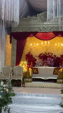 This media file depicts a decorated stage set for an event, likely a wedding reception or a similar celebration, in Keudematangkuli, Indonesia. The stage is elaborately adorned with a golden backdrop, featuring ornate carvings and a rich maroon curtain draping across the top. A plush white sofa sits centrally on the stage, flanked by two elegant, light-colored armchairs. A large floral arrangement, bursting with red and pink blooms, is positioned behind the sofa, adding a vibrant splash of color. Smaller floral arrangements and greenery are placed on either side of the stage, near the armchairs, and at the base of a tiered platform in front of the stage, which is covered in white fabric.

Overhead, cascading crystal-like decorations add a touch of glamour. The lighting appears to be warm and ambient, with chandeliers and fairy lights illuminating the scene, suggesting the event is taking place in the evening or indoors. To the right of the stage, a black jacket is draped over a stand, and an orange plastic chair is visible in the foreground, with another orange chair and a black fan further in the background, hinting at the backstage or preparation area. The overall impression is one of a formal and festive occasion, with a focus on elegance and beauty. There is no visible text in the media file.