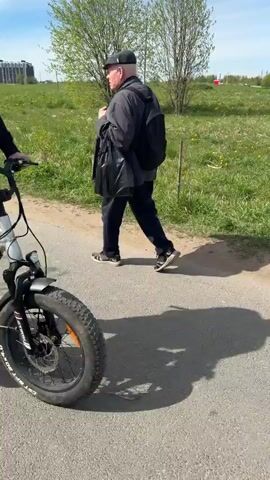 The video shows a person walking alongside an electric bike on an asphalt path, with a grassy field and buildings in the background.  The camera then pans, revealing a man pushing a baby stroller on the same path. Two individuals are riding electric bikes ahead of them, moving away from the camera. Overhead electrical wires are visible along the side of the path, and apartment buildings line the edge of the frame.