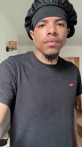A young, light-skinned man stands indoors. He is wearing a black t-shirt with a small red Levi’s logo and a black satin bonnet. He has facial hair, a tattoo on his neck, and what appears to be an earring on his left ear. He holds a long, white and black extendable rod, extending it in both hands, then twirling it. The interior space appears to be an apartment. Behind him are a television, a rattan wall lamp, a dining table with chairs, and another room. Loud music plays.