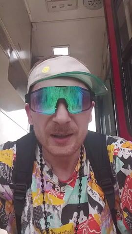 The video shows a close-up of a man wearing a colorful shirt, a black backpack, and two necklaces. He also has a white visor with a green brim, and large iridescent sunglasses that reflect the interior environment. He appears to be inside some type of vehicle, perhaps a train or bus, and is speaking into the camera. The interior of the vehicle is visible in the background, with overhead lights and other details suggesting a public transportation setting. The man has light skin, reddish hair, and a trimmed mustache and beard. His facial expression is animated as he speaks.