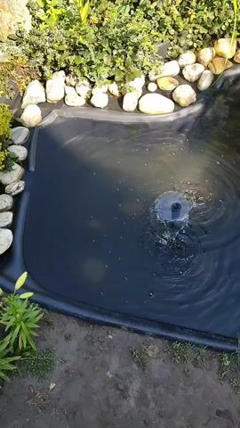 This media file shows a small, black-lined garden pond with a circular fountain in the center. The water in the pond is dark and rippled from the fountain's spray. The pond is surrounded by a border of smooth, light-colored stones and lush green foliage. Some yellow-green plants with long leaves are visible at the bottom left of the frame. The ground around the pond is dirt with patches of small green weeds. The scene appears to be outdoors, likely in a garden, with shadows suggesting it is daytime. There is no visible text in the media file.