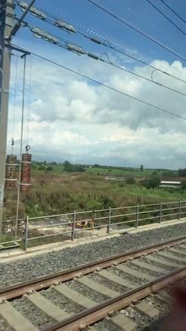 The video presents a view from a moving train, looking out over a rural landscape. In the immediate foreground, the train tracks, composed of dark sleepers and grey ballast, are prominently visible, parallel to the direction of travel. A low, grey metal fence runs alongside the tracks, separating them from the broader landscape.

Beyond the fence, the scene unfolds with expansive green and brownish fields, some appearing cultivated in rows, while others are wilder and overgrown with tall grasses. A small, narrow body of water, possibly a river or canal, briefly appears in the midground, reflecting the sky. Scattered trees and small clumps of foliage dot the landscape, some appearing green and lush, while others show drier, more subdued tones.

Throughout the view, multiple large metal power line pylons stand tall, stretching across the fields and into the distance, with numerous overhead power lines crisscrossing the sky. In some parts of the fields, bright orange or red netting or tarp is visible, possibly indicating agricultural activity or construction zones.

The sky above is a mix of bright blue patches and large, fluffy white clouds, suggesting a partly cloudy but generally sunny day. Distant hills or mountains can be faintly discerned on the horizon, blending with the sky. The continuous, steady motion indicates the train is traveling at a consistent speed through this pastoral Italian landscape near Teverola.