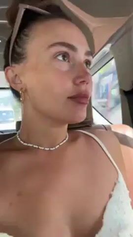 A woman is sitting in the back seat of a moving car. She is wearing a white top and a beaded necklace, and her hair is in a bun with sunglasses on top of her head. She is looking directly at the camera with a slight smile.  The background shows glimpses of the outside environment through the car window.