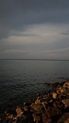 This is a vertical video of a body of water, likely a lake or ocean, with a rocky shoreline in the foreground. The sky transitions from dark, cloudy conditions to a bright, sunny sky with the sun low on the horizon, indicating either sunrise or sunset. The water appears calm with small ripples. The lighting changes from dim to bright as the sun becomes more prominent. No people or specific objects are clearly visible beyond the natural elements.