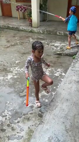 This media file captures a playful scene in Dujiangyan City, China, likely during daytime. The setting appears to be an outdoor courtyard or patio with a wet, stained concrete floor, indicating recent water activity. Sunlight is visible, suggesting it is daytime.

The primary subjects are two young children and two adults. In the foreground, a young girl in a patterned swimsuit and light pink sandals is holding a yellow and orange toy water gun. Her hair is wet, and she looks towards a camera being held by an adult. She appears engaged and possibly amused.

In the background, another child, wearing a blue shirt, blue swim trunks, and a blue cap, is visible. This child is also holding a toy water gun and appears to be spraying water. Two adults are present in the background; one is holding a smartphone and observing, while another is holding a blue toy water gun.

The activity taking place is a water gun fight or playful water play. The wet environment and the presence of water guns strongly suggest this. The overall mood seems lighthearted and joyful, especially from the expression of the young girl in the foreground. There is no visible text in the media file.