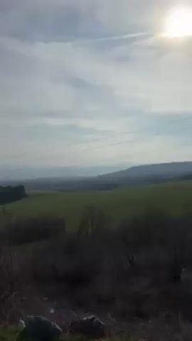 The video shows a landscape view on a slightly overcast day. In the foreground, there are bare bushes and some rocks. In the midground, there is a green field interspersed with darker patches of trees. In the background, there are hazy mountains. Power lines stretch across the top of the scene. The camera is stationary.
