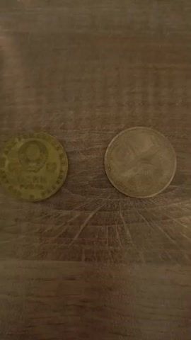 The video shows two coins lying on a wooden surface. The coin on the left is round and appears to have some writing and a symbol in the center. The coin on the right is also round and has an image on it. The lighting is dim, and the surface has a visible wood grain. The video is brief and does not show any people or actions.