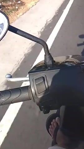 The video shows a first-person perspective of someone riding a scooter on a road. The road has a white line marking the side of the lane. To the left of the road, there is a sidewalk with some houses and trees. The scooter's handlebars and instrument panel are visible, including a turn signal switch and a mirror. The rider's feet, wearing sandals, are also visible, as is the shadow of the rider and the scooter on the road. The overall environment appears to be a sunny day in a suburban or rural area.