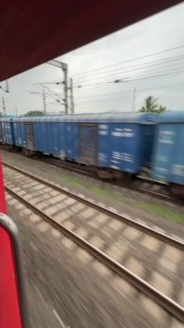 A first-person perspective shot from a moving train shows another train passing by on an adjacent track. The passing train consists of several blue, boxcar-style freight cars. The sky is overcast with clouds, and utility poles and wires are visible in the background. The foreground is dominated by the red interior of the train the viewer is on, and the blurred motion of the train tracks.