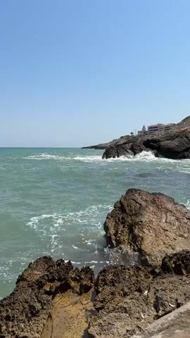 The video displays a view of the ocean with waves crashing against rocks. The water is a greenish-blue color, and the sky is clear and blue. In the background, there is a rocky coastline with some buildings visible on top of the cliff. The foreground features large, rough-textured rocks with some water splashing over them. The overall scene is one of a sunny day at the coast.