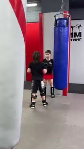 Two young boys in boxing gear stand facing each other in a gym. One boy wears black shorts and a black t-shirt, with shin guards and gloves. The other boy wears red shorts and a black t-shirt, with shin guards and gloves. A large red and white punching bag stands to the left of the frame. A blue punching bag hangs behind the boys. The floor is gray with a tiled pattern. A sign with the letters "MM" is visible in the background. One boy speaks to the other as they stand ready to box.