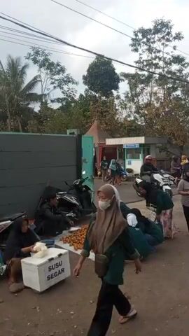 The video shows an outdoor scene, possibly a street market or a gathering area, with several people and motorcycles. In the foreground, a group of individuals, mostly women wearing headscarves, are gathered around a large pile of brown, spherical objects, likely fruit or vegetables, spread on a white cloth on the ground. Some people are seated while others are standing. A black motorcycle is parked to the left of the pile. Further back, there are more people, some on motorcycles, and small stalls or structures with awnings. One structure has signage that appears to read "SATPAM." The overall atmosphere suggests a busy, public space with commercial activity.