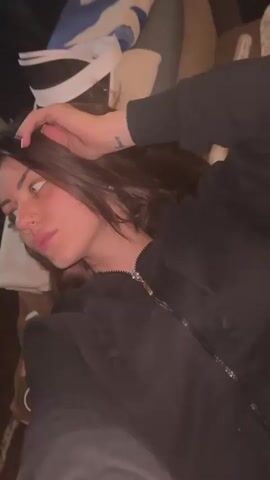 A young woman with dark hair and freckles lies down in an indoor setting, possibly on a couch or bed. She wears a black zippered hoodie and has a nose piercing and a small tattoo on her right wrist.

At the beginning of the video, she is seen lying with her head turned to the left, gazing off-camera with a neutral or slightly pensive expression, her right hand resting near her head. After a couple of seconds, she turns her head to face the camera, revealing a relaxed and pleasant smile. She briefly closes her eyes while maintaining the smile. The video concludes with a quick, blurry motion, suggesting an abrupt end or a sudden camera movement.

The background is somewhat cluttered with various indistinct objects, including what appears to be white and blue fabric, a white visor-like item, and a white tube or rolled-up paper. The lighting is soft, indicating an indoor environment, possibly in the evening or under ambient artificial light. No visible text is present.