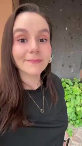 A woman is recording a video of herself. She is wearing a black shirt and has long brown hair and gold earrings. She is also wearing a gold necklace with a clover pendant. Her makeup includes pink blush and eyeshadow. Behind her is a white car and what appears to be the outside of a house. Another person's arm holding a black object moves in the frame. The woman smiles at the camera throughout the video.
