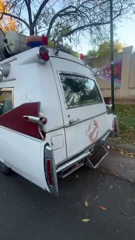 The camera pans across the side and back of a white Ghostbusters-themed vehicle. The car has a "Ghostbusters" logo and the words "Ecto1 Argentina" on its side. Additional gadgets and lights are mounted on the roof. The car is parked on a street with trees and a mural in the background.