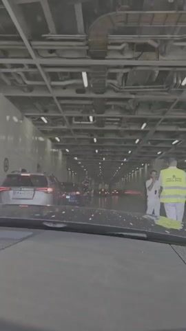The video shows a view from inside a car, looking forward through the windshield. The car appears to be on a ferry, as evidenced by the enclosed, industrial-looking space with a low ceiling, exposed pipes and conduits, and overhead lighting. The car is in a lane of other vehicles, mostly SUVs and sedans, also moving forward. On the right side of the frame, two individuals in uniform are visible. The person closer to the camera is wearing a light-colored shirt and pants and is gesturing forward with their right hand. The second person, slightly further away, is wearing a high-visibility yellow vest over a light-colored uniform and appears to be standing behind the first person. The overall environment is dimly lit, with the primary light sources appearing to be the overhead lights and the brake lights of the vehicles ahead. The perspective suggests the car is in motion, slowly proceeding through the ferry's car deck.