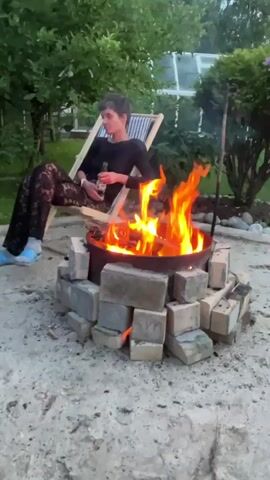 A person sits in a striped chair by a campfire. The person is wearing black lace pants and a black shirt. They are holding a bottle of beer. The campfire is in a metal ring surrounded by bricks. There are trees and a greenhouse in the background. The ground is covered in gravel.