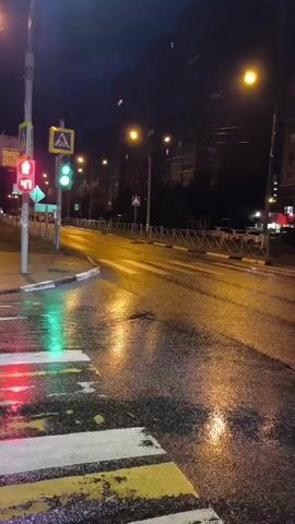 The video shows a wet street at night, likely after rain. A traffic light is visible, with the red light illuminated for the direction facing the camera and the green light illuminated for the perpendicular direction. A pedestrian crossing is marked with white and yellow stripes on the wet asphalt. Streetlights cast reflections on the wet road surface. In the distance, there are buildings and more streetlights. There are no people clearly visible in the frame. The scene appears to be a quiet intersection.