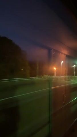 The video shows the view from the window of a moving train at night. Outside, the blurred landscape features streetlights and a train platform. The interior of the train is visible, with reflections on the window and a panelled wall. The overall lighting is dim, with the exterior lights providing the main illumination. The camera appears to be handheld, and the video is somewhat shaky due to the train's motion.