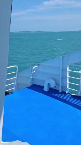 The video pans across the deck of a ferry, showing the bright blue deck with white railings. In the background, the turquoise ocean stretches to the horizon under a partly cloudy blue sky. Distant islands are visible on the horizon. A life preserver is attached to the railing in the mid-ground. The camera is positioned on the ferry, looking out towards the open sea.