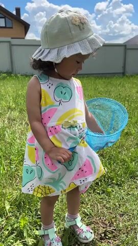 A young child wearing a white hat with a floral decoration and a white sleeveless dress with a fruit pattern stands in a grassy area. The child holds a blue butterfly net in their right hand. A house and a white fence are visible in the background. The sky is blue with white clouds. The child is looking down and to the right, then turns their head to look directly at the camera.