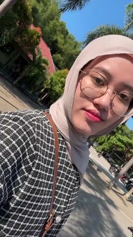 A young woman wearing a hijab and glasses smiles and laughs directly into the camera in a selfie-style video. She has bright pink lipstick on and appears to be outdoors in a sunny environment. She is wearing a black and white patterned jacket. The background shows trees, a building, and a paved walkway.