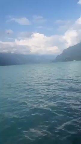 The video shows a view of a lake with mountains in the background. The water is a light blue color and has small ripples on the surface. The sky is blue with scattered white clouds. The mountains in the background are partially obscured by clouds.