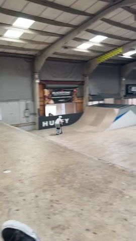 Here is a detailed description of the video:

A person wearing a white shirt, light blue jeans, white shoes, a white helmet, and knee pads is riding a scooter in an indoor skate park. The person rides up a curved ramp, jumps high in the air, rotates, and lands on a second ramp. The ramps are made of wood and painted with different colors. The indoor skate park has high ceilings with metal beams, gray walls, and various advertisements. There is a "HUFFY" sign near the ramp and a "BLUNT" banner in the background.