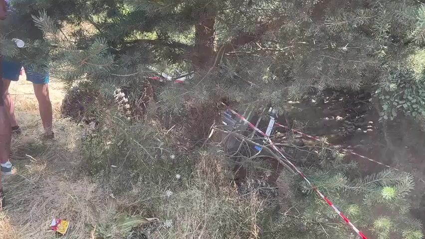 The video shows a downed off-road vehicle partially obscured by a pine tree and underbrush. The vehicle appears to be on its side. Several people are standing to the left of the frame, observing the vehicle. Red and white caution tape is visible, indicating a restricted area. The environment is outdoors, with dry grass and foliage. A stream or body of water is visible in the background.